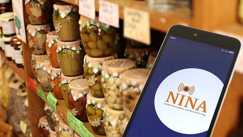 Regal mit Lebensmitteln im Hintergrund und Handy mit NINA-App im Vordergrund